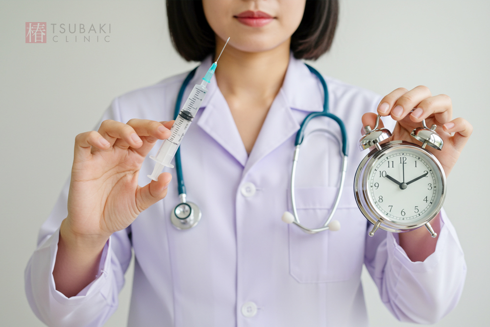 白衣を着た女性が、右手に注射器を、左手に時計をもって立っている画像。