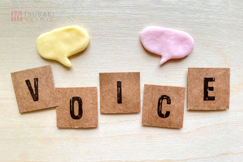 木目の背景の上に、紙の文字で「VOICE」と並べられています。上には黄色とピンクの吹き出し形の飾りがあり、体験談や口コミを紹介するイメージです。