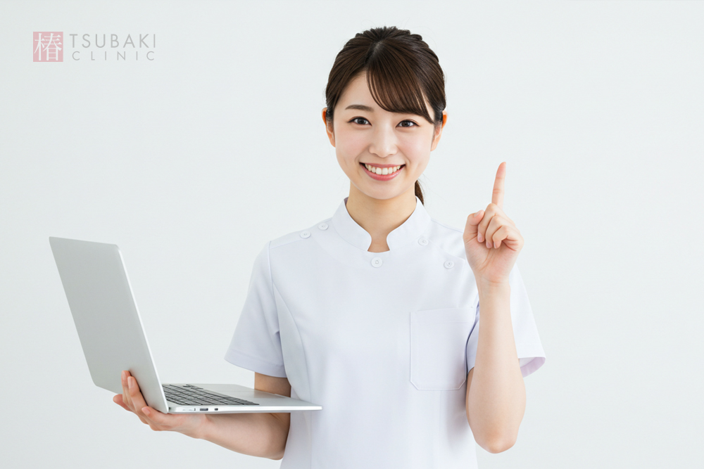 30代くらいの女性スタッフが白い制服を着て、ノートパソコンを持ちながら人さし指を立ててポイントを説明しています。やさしく笑顔でこちらを見ている、クリニック案内のイメージです。