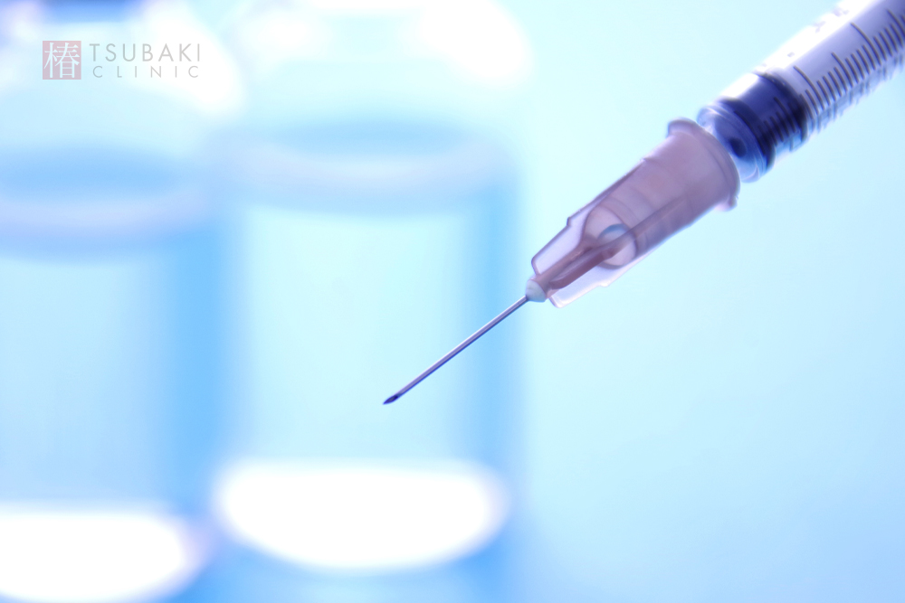 青みがかった背景の前で注射器の針先がアップで写されており、背後にぼかした薬剤バイアルが並ぶ、脂肪溶解注射などセルライト治療をイメージさせる医療シーン