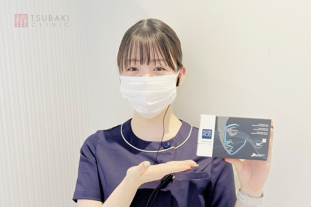 椿クリニックの院内で、ネイビーの制服を着たマスク姿のスタッフが「スネコスパフォルマ」の箱を手に持ち、もう一方の手で商品を紹介している様子。